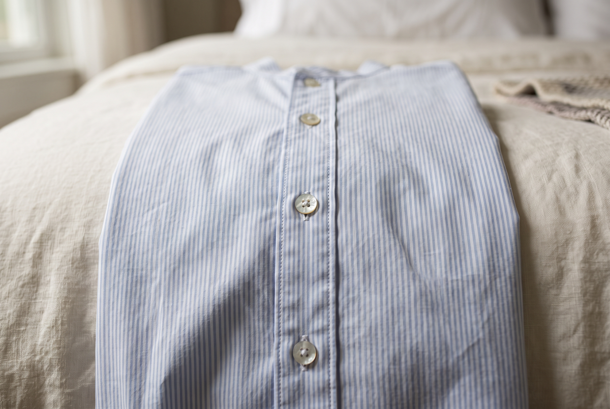 Gros plan sur la patte boutonnée d'une chemise de nuit en popeline rayée bleu pâle, une main fermant un bouton nacré Image